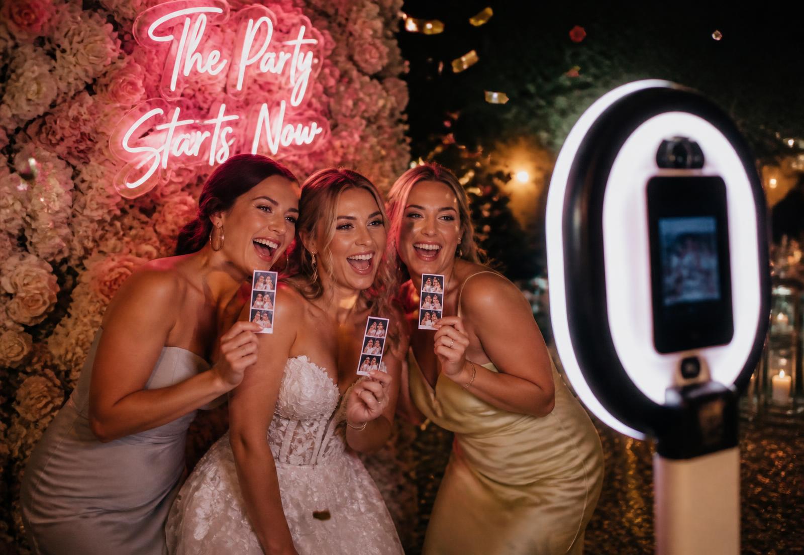 Guests laughing in an open-air photo booth holding printed photo strips at a wedding